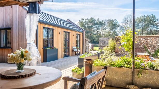 A wooden deck with a round table and chairs, a fan, and a view of a house with a metal roof and wooden siding. - The Able Stables - Sam's Cottage, East Rudham, Norfolk (East Rudham)
