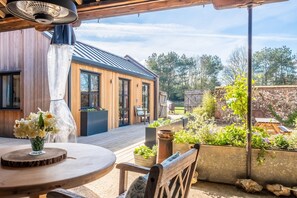 Een houten terras met een ronde tafel en stoelen, een ventilator en uitzicht op een huis met een metalen dak en houten bekleding.