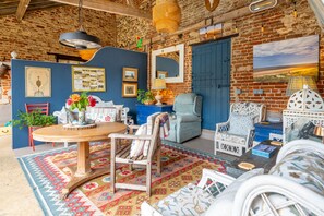 A rustic interior with exposed brick walls, a round wooden table, and a variety of seating options including a blue armchair and a white patterned chair. - The Able Stables - Sam's Cottage, East Rudham, Norfolk (East Rudham)