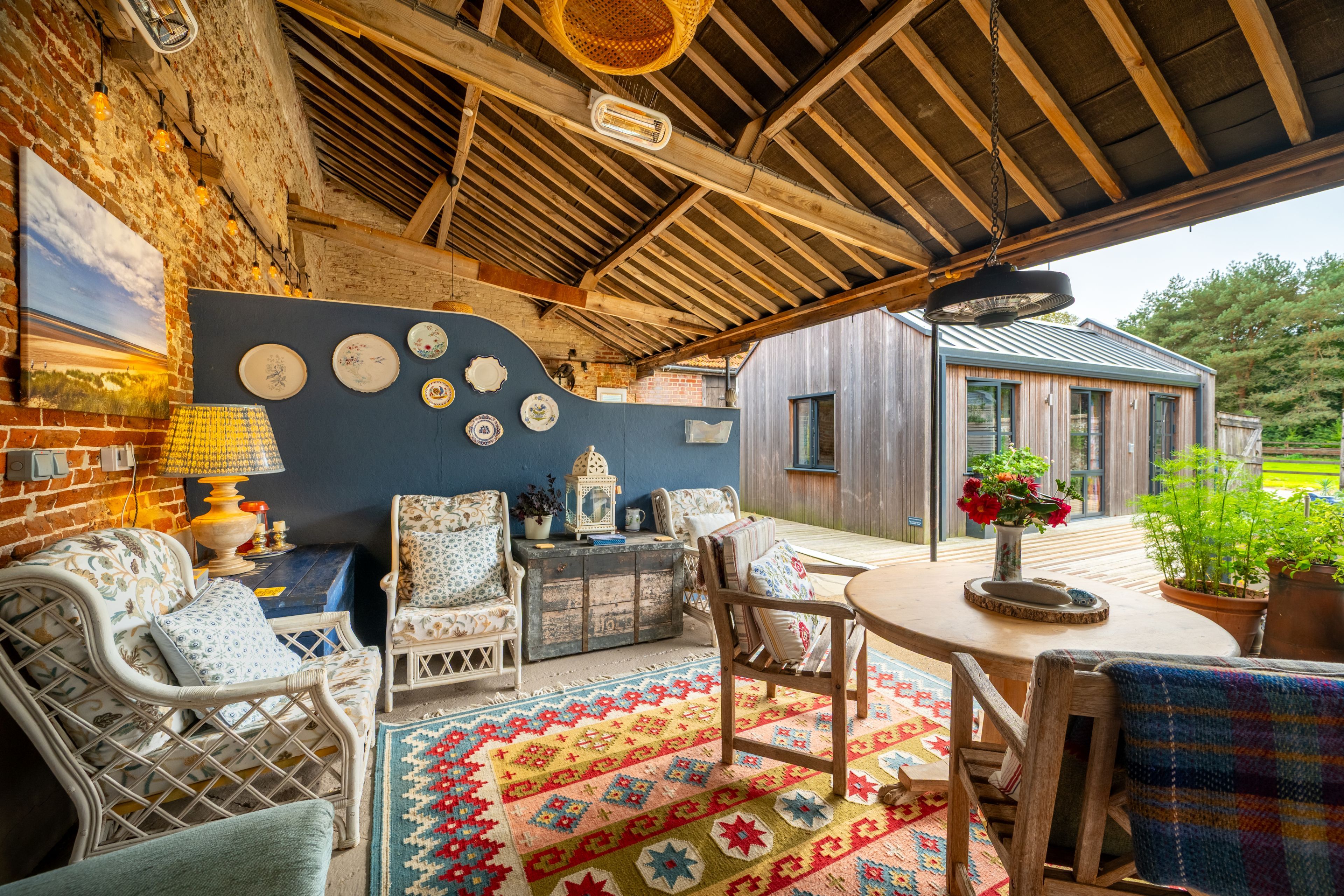 A covered outdoor seating area with wicker furniture, a patterned rug, and a view of a wooden building.