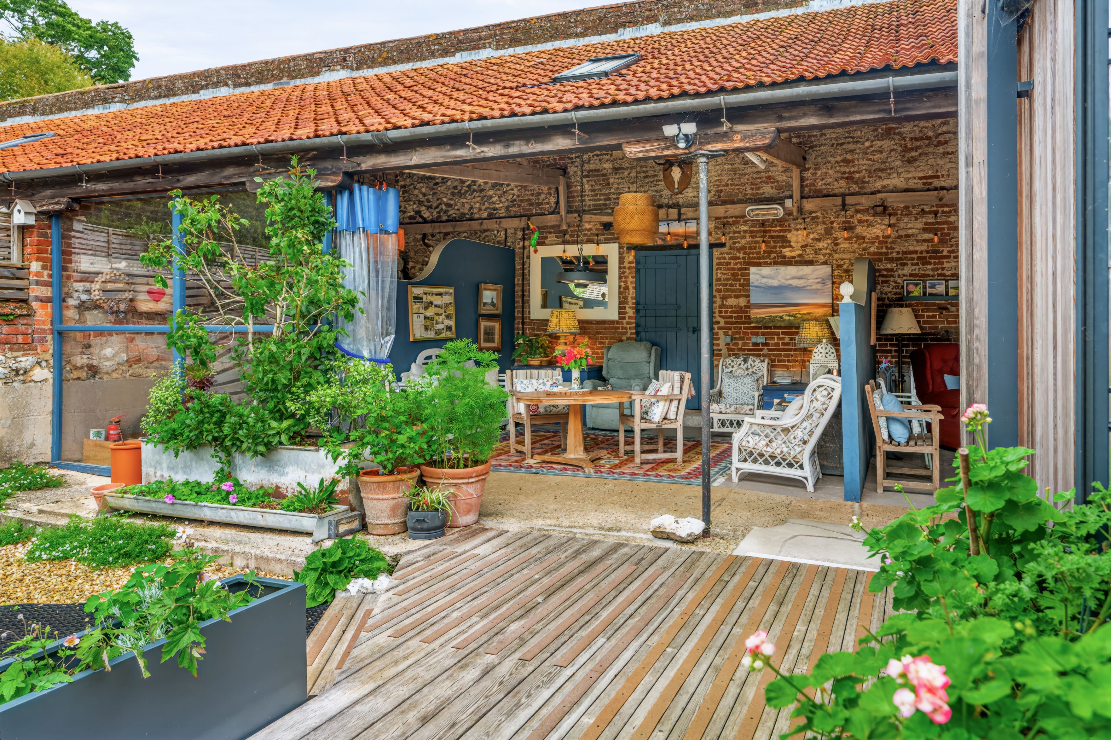 A covered outdoor area with a wooden deck, potted plants, and a brick wall.