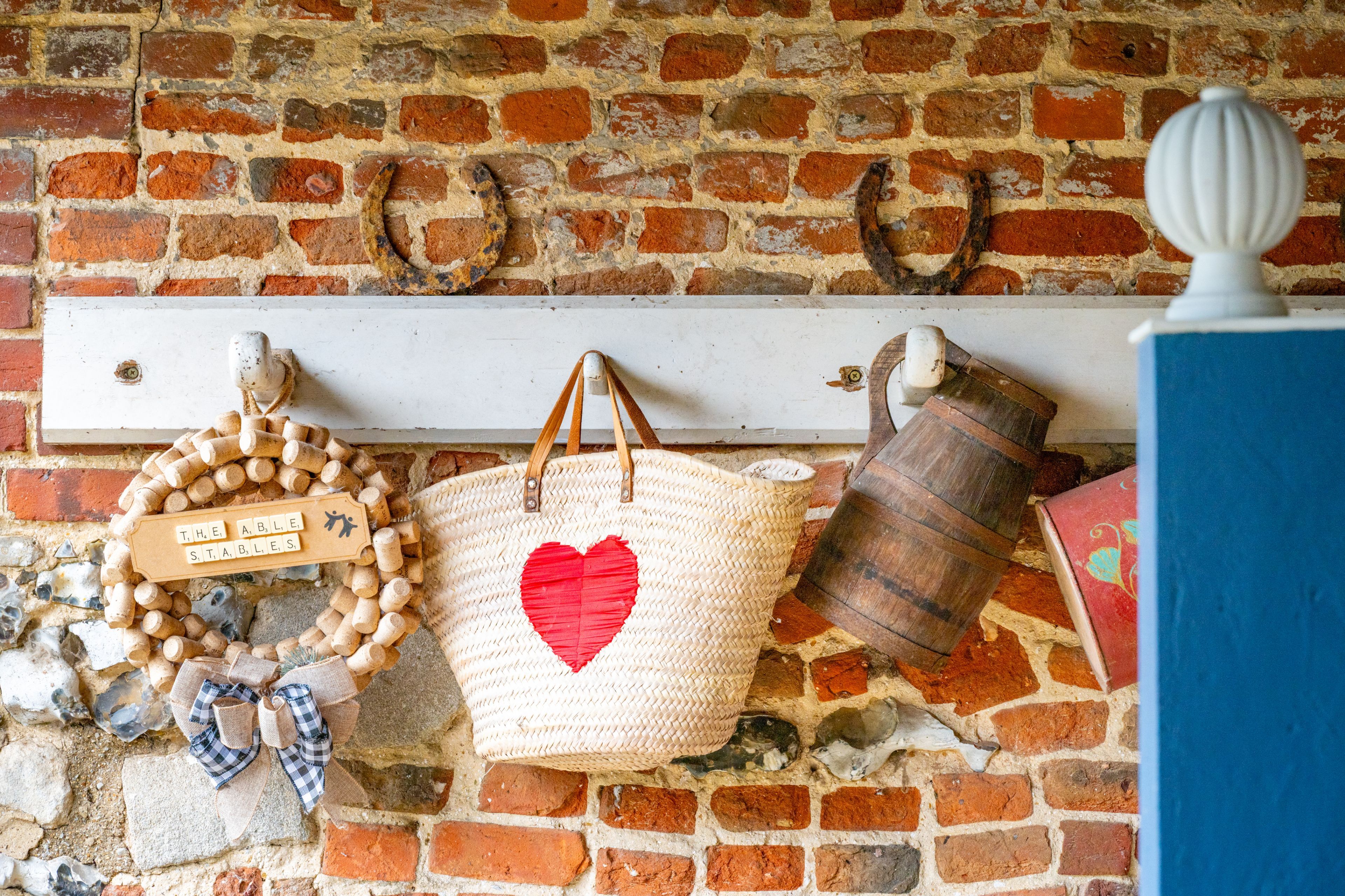 A rustic brick wall with a white shelf holding a woven basket with a heart design, a wooden bucket, and a decorative wreath.
