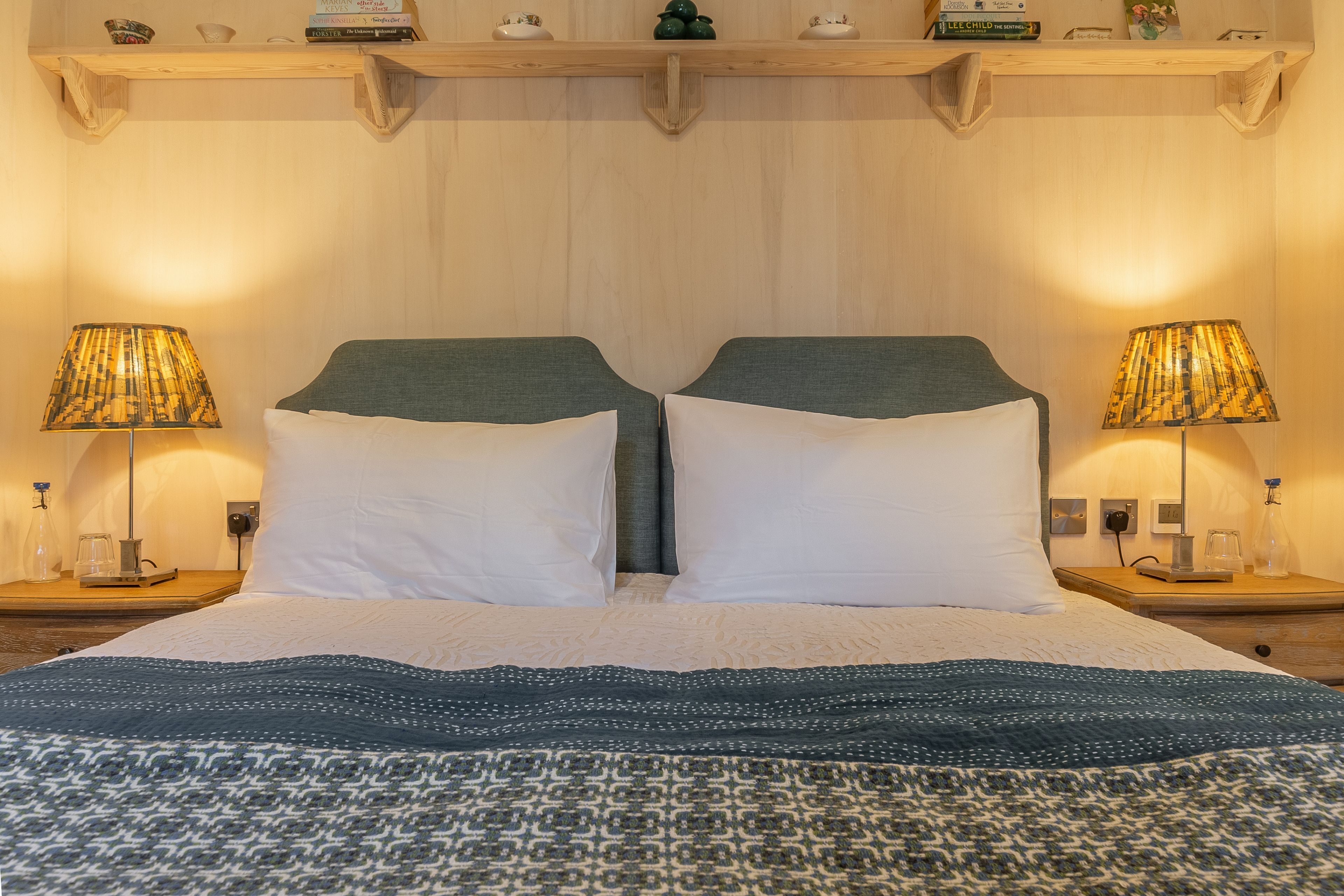 A double bed with a patterned bedspread, two bedside lamps, and a wooden shelf above the bed.