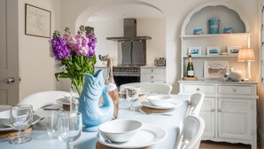 Un coin repas avec une table blanche dressée pour quatre personnes, un vase rempli de fleurs violettes et, au fond, une cuisine.