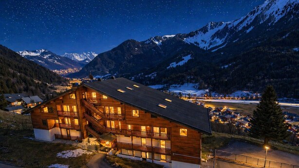 Eine Holzchalet mit mehreren Balkonen, einer Treppe und Blick auf schneebedeckte Berge unter einem sternenübersäten Nachthimmel.