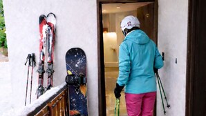 Eine Person in Skiklamotten steht vor einer Tür, neben der Ski und ein Snowboard an der Wand lehnen.