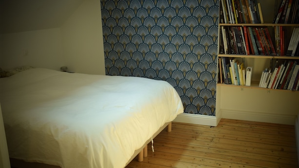 Een slaapkamer met een bed, houten vloeren, een boekenplank en behang met een patroon.