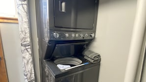 A compact, wall-mounted washer and dryer unit in a small room with wooden flooring and a curtain. - Water Front Vacation Home (Clay Township)