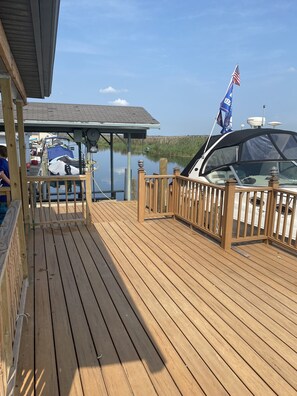 A wooden deck with a covered area, a boat docked with a flag, and a calm water body in the background.