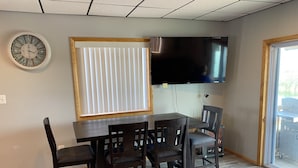 A dining area with a table and chairs, a clock, a TV, and a window with blinds. - Water Front Vacation Home (Clay Township)