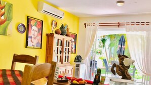 A room with a yellow wall, a wooden chair, a table with a glass top, and a window with a view of palm trees. - 2 connected villas 19 Pax (Playa del Carmen)
