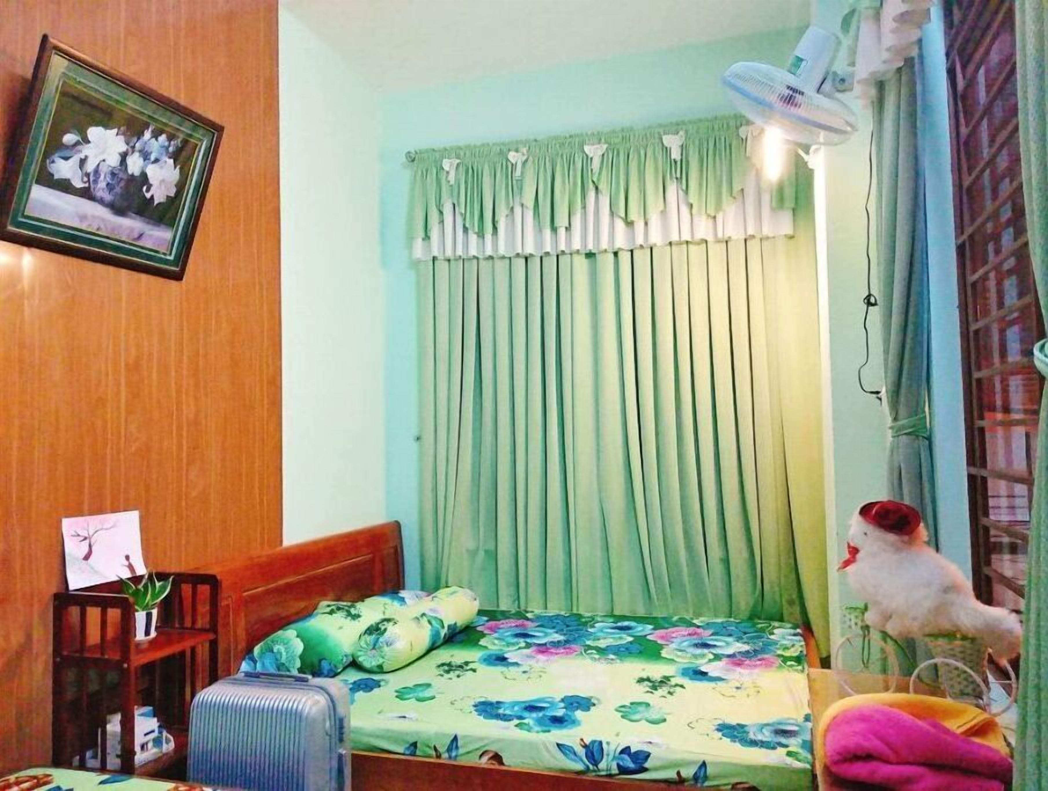 Een slaapkamer met een bed, nachtkastje, stoel, koffer, ventilator en raam met gordijnen.