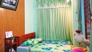 Une chambre à coucher avec un lit, une table de chevet, une chaise, une valise, un ventilateur et une fenêtre avec des rideaux.