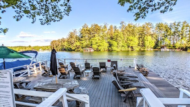 Une terrasse en bois avec des chaises longues et des tables, donnant sur un lac et entourée d’arbres.