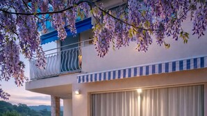 Un espace de restauration en plein air avec une table dressée pour deux personnes, entourée de chaises blanches, sous un balcon orné de fleurs violettes.