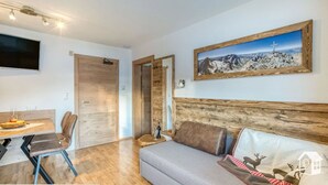 Ett modernt vardagsrum med en sänggavel i trä, en grå soffa, ett matbord med stolar och en väggmonterad TV.