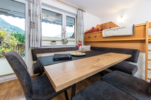 A room with a wooden table, grey upholstered chairs, a window with curtains, a bed with a headboard, and a view of mountains.