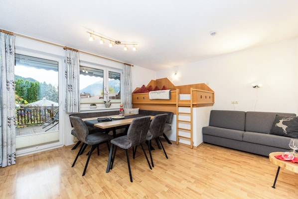 Une chambre moderne comprenant une table à manger, des chaises, un canapé, un lit mezzanine en bois et un balcon offrant une vue sur les montagnes.