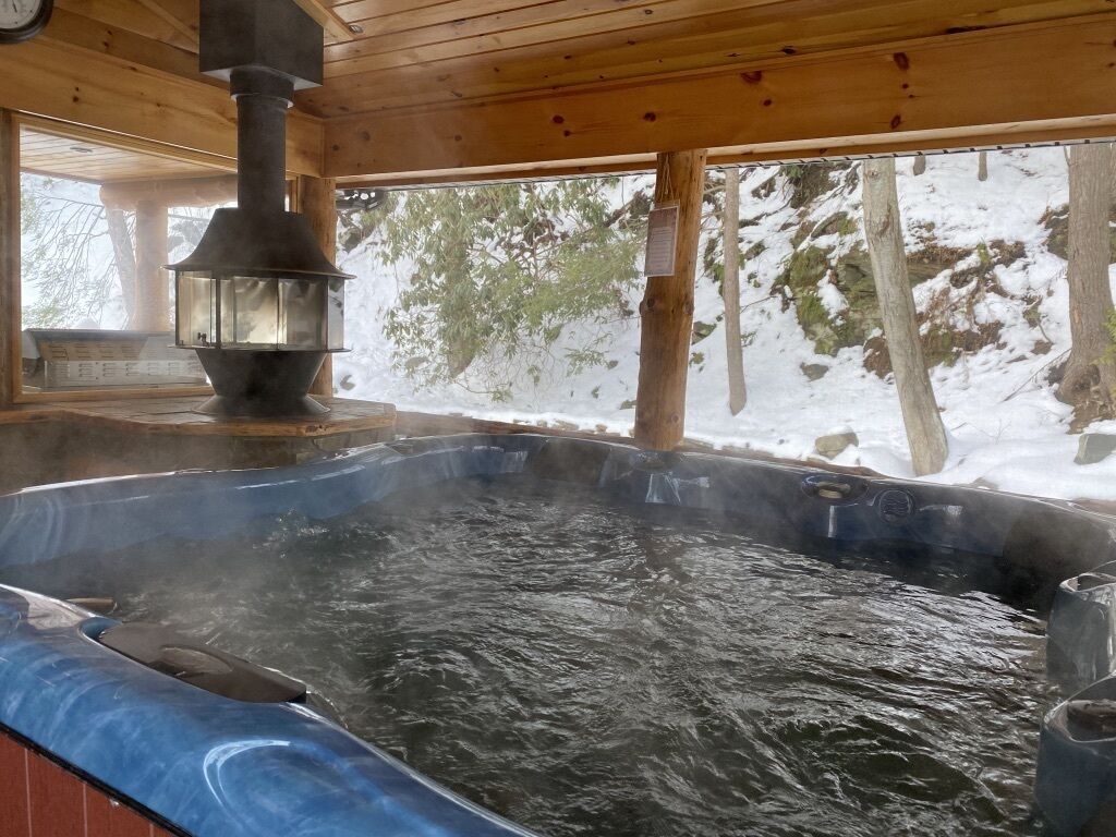 A hot tub in a wooden cabin with a wood-burning stove above it.