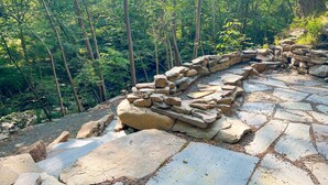 Un sendero de piedra en un área boscosa, con un muro de piedra y árboles al fondo.