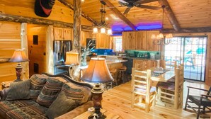Interior rústico de una cabaña con techo de madera, comedor, cocina y sala de estar con sofá y lámparas.