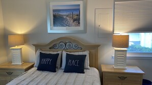Ein Schlafzimmer mit einem Bett, zwei Nachttischen mit Lampen, einem Schminktisch, einer Kommode, einem gerahmten Bild an der Wand und einem Fenster mit Jalousien.