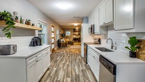 A modern kitchen with white cabinets, a dishwasher, and a microwave. There is a wooden floor and a window with a view of the living room.