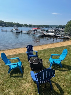 A lakeside dock with several boats, a grassy area with blue Adirondack chairs, and a round table.