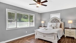 Une chambre à coucher avec un grand lit, deux tables de chevet, un ventilateur de plafond et une fenêtre avec des stores.