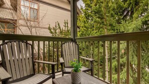 Un espace extérieur aménagé avec une zone de repos couverte, comprenant deux fauteuils Adirondack et une petite table, le tout entouré d’une balustrade en bois et offrant une vue sur une maison et des arbres.