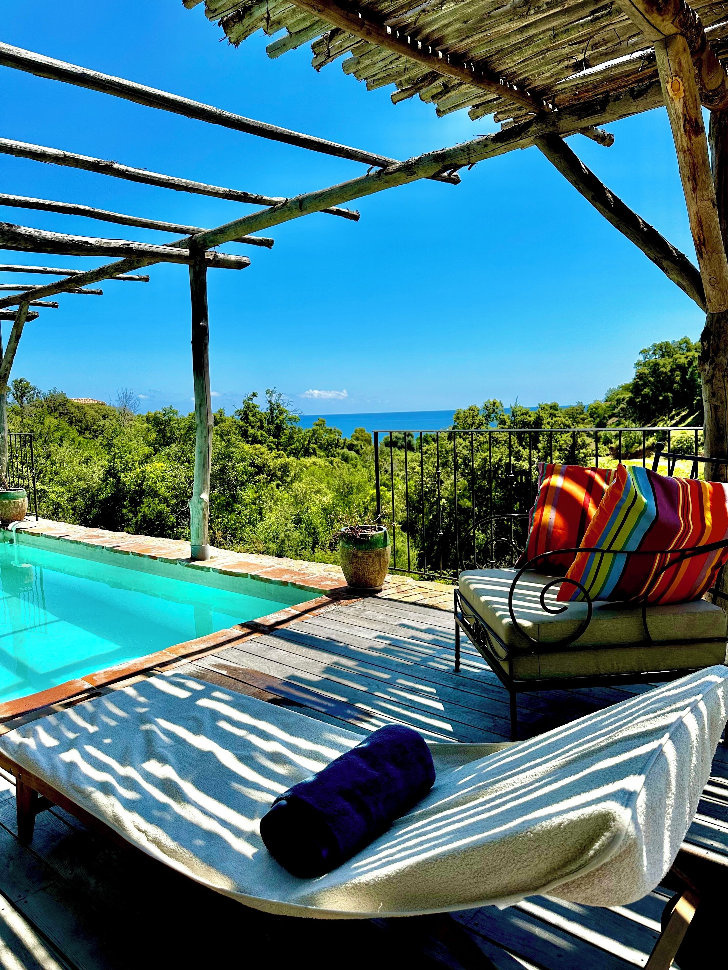 Un espace aménagé au bord de la piscine, avec un fauteuil de relaxation et un coussin coloré, une pergola en bois et une vue sur la mer.