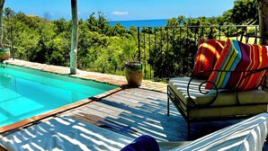 Zona junto a la piscina con una silla de descanso y un cojín colorido, una pérgola de madera y vistas al mar.