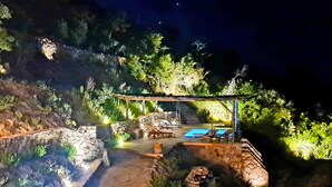 Un espacio al aire libre de noche con un muro de piedra, una pérgola de madera y una piscina.