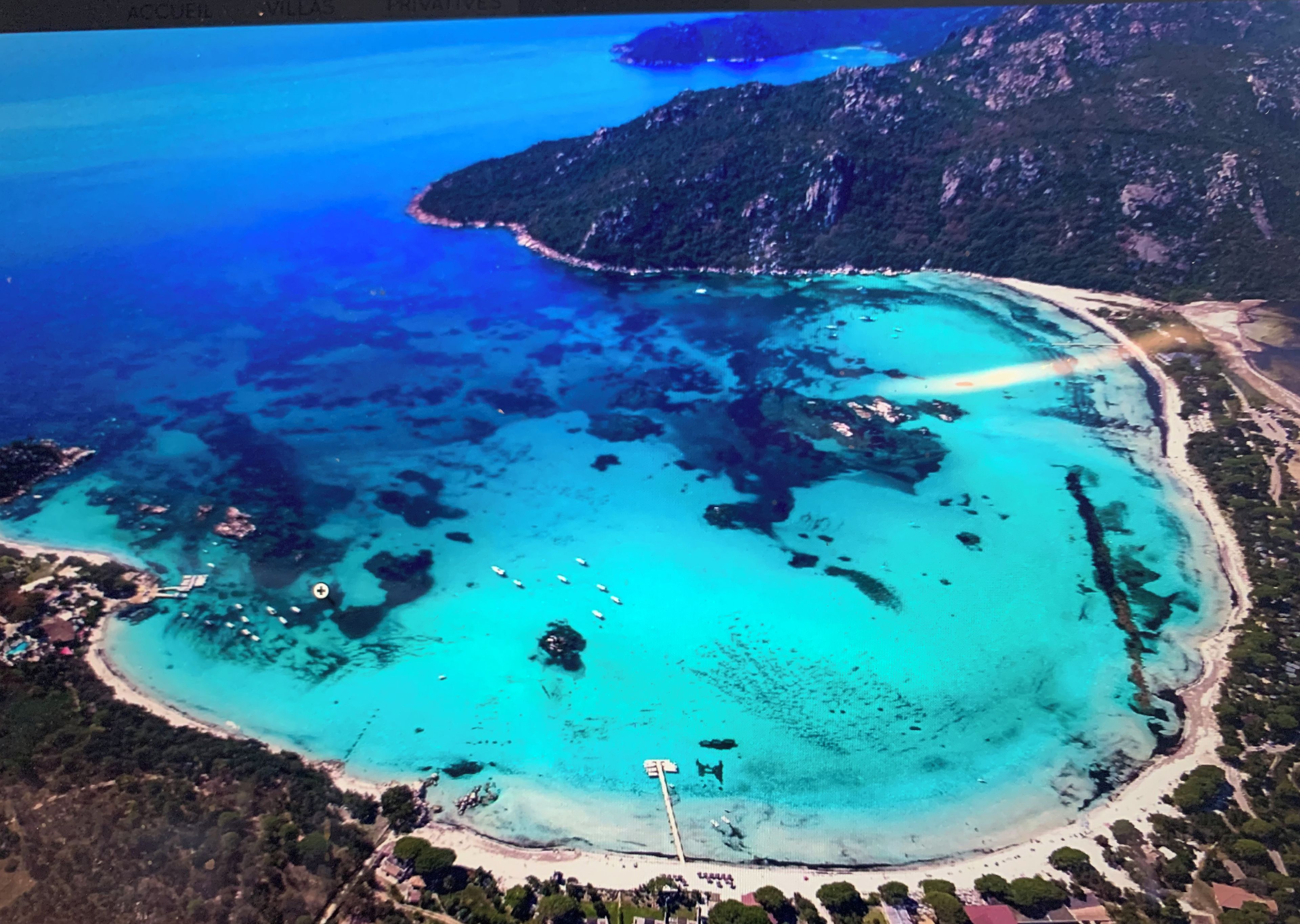 An aerial view of a coastal area with clear turquoise waters, a sandy beach, and a mountainous backdrop.