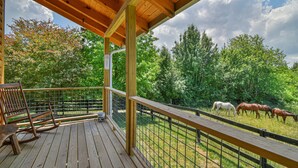 Eine Holzterrasse mit Geländer, einem Schaukelstuhl und Blick auf Pferde, die auf einer Wiese weiden.