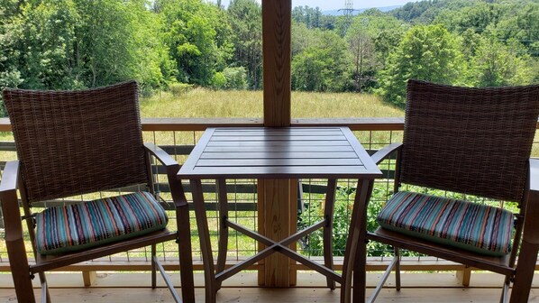 Una terraza de madera con dos sillas de mimbre y una mesita pequeña, con vistas a un paisaje verde y exuberante.