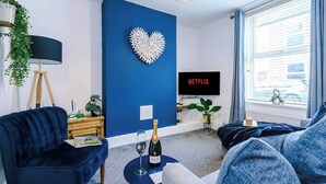 Ett modernt vardagsrum med en blå accentvägg, en hjärtformad dekoration, en tv-skärm som visar ”NETFLIX” och ett fönster med persienner.