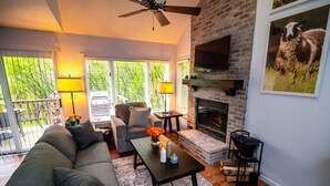 Ein Wohnzimmer mit einer Couch, einem Couchtisch und einem Kamin. An der Wand ist ein Fernseher angebracht und es gibt einen Balkon mit Geländer.