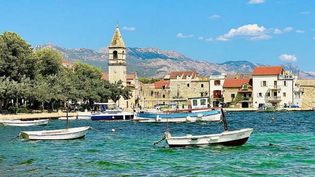 Uma vila costeira com uma torre de igreja imponente, barcos atracados ao longo da orla e edifícios com telhados de terracota.