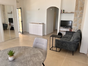 Een moderne woonkamer met een grijze bank, een klein rond tafeltje met een plant, een wit kastje en een tv die aan een stenen muur is bevestigd.