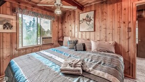 A wooden cabin bedroom with a bed, ceiling fan, large window, and artwork.