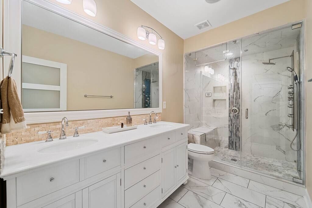 A modern bathroom with a large mirror, two sinks, a toilet, and a glass-enclosed shower.