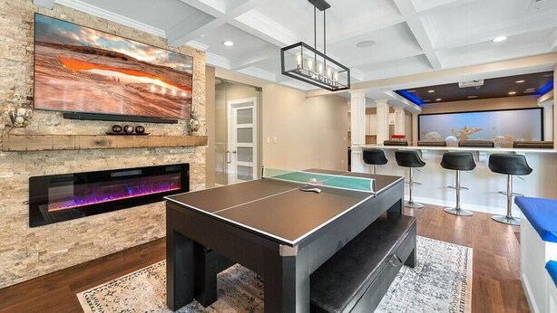 A modern living room with a ping pong table, a fireplace, a large TV, and a bar area with stools.