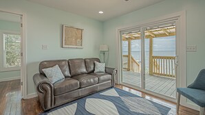 A living room with a brown leather couch, a blue rug, and a view of a wooden deck and water. - Sunsets and Relaxation await -  Spot of Sun Bay Cottage (Gulf Shores)