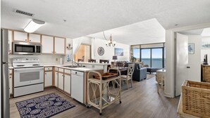 A modern kitchen with white cabinetry, a microwave, and a stove. There is a dining area with a table and chairs, and a view of the ocean through large windows.