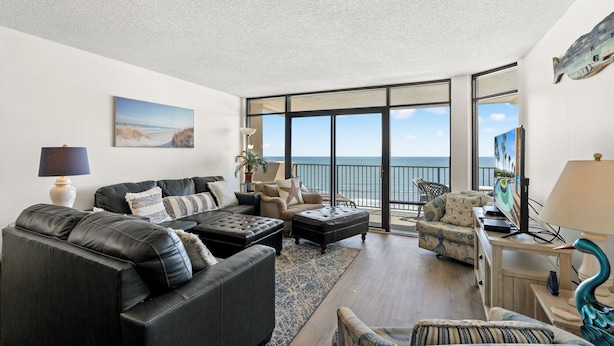 A spacious living room with a large window, a black leather sofa, a wooden coffee table, and a small side table with a lamp.