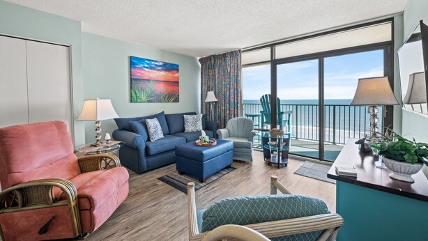 A spacious living room with a blue sofa, a pink armchair, and a teal armchair. There is a large window offering a view of the ocean.