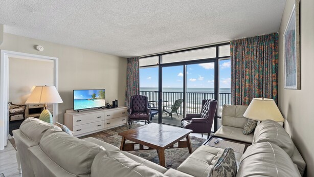 Un salon avec un canapé, une table basse et un téléviseur, un balcon avec des chaises et une vue sur l’océan.