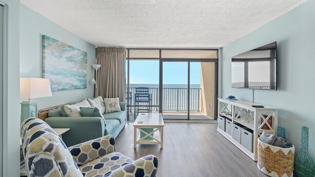 A living room with a sofa, armchair, and coffee table, a TV mounted on the wall, and a balcony with a view. - Verandas 1102 - 1 Bedroom Ocean Front Condo (North Myrtle Beach)