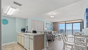 A modern kitchen with a central island, a dining area with white chairs, and a large window offering a view of the sky. - Verandas 1102 - 1 Bedroom Ocean Front Condo (North Myrtle Beach)